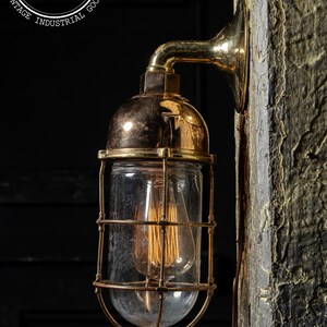 May include: A brass industrial wall light with a clear glass shade and a caged bulb. The light is mounted on a rough textured wall.