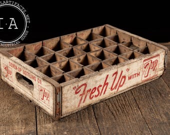Op de afbeelding: Vintage houten 7-Up frisdrankkrat met meerdere compartimenten. De krat heeft vervaagde rode letters met de tekst "Fresh Up with 7-Up" en "Seven-Up Bottling Co." aan de zijkant. De krat is gemaakt van verweerd hout en staat op een houten oppervlak.