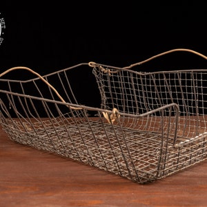 May include: A vintage wire basket with two leather handles. The basket is made of a wire mesh and has a rectangular shape. The basket is sitting on a wooden surface.