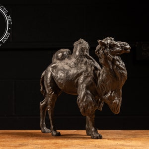 May include: A bronze sculpture of a camel standing on a wooden surface. The camel is facing to the right and has a hump on its back. The sculpture is detailed and realistic.
