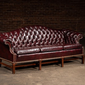 May include: A burgundy leather Chesterfield sofa with a button-tufted back and rolled arms. The three-seat sofa has a wooden frame with brass nailhead trim and is set against a brick wall.