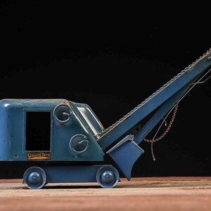 May include: A vintage blue metal toy crane featuring a long boom arm with a metal chain and a small bucket attachment. The side of the cab displays a rectangular black and gold label with the text "STRUCTO TOYS" in a clear, legible font.