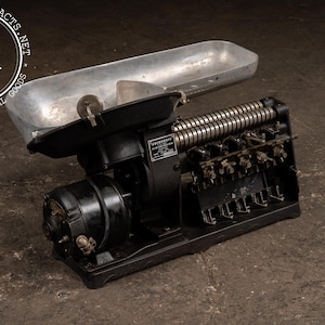 May include: Vintage industrial scale with a black base, silver tray, and a series of metal coils. The scale has a label that reads "CLEVELAND". The background is a dark, textured surface.