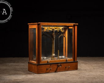 Puede incluir: Balanza antigua de madera con una caja de vidrio y detalles en latón. La balanza tiene dos plataformas y una viga central con pesas. La balanza está en buen estado y tiene una estética vintage.