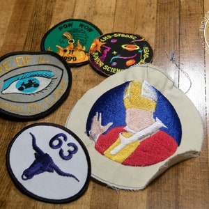 May include: Five embroidered patches with various designs. The patches are circular and feature a blue eye with a motorcycle, a blue and white bull's head, a yellow and red figure with a yellow hat, a green patch with a Native American figure, and a black and white patch with a sun and planets.