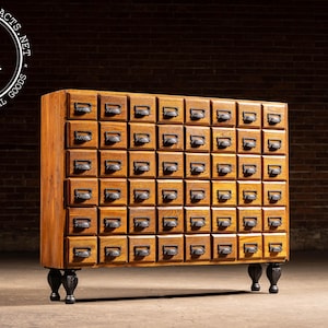May include: A vintage wooden cabinet with numerous small drawers, each with a metal pull. The cabinet is made of light brown wood and stands on four black, ornate feet. The background is a brick wall.
