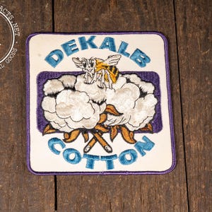 May include: Vintage Dekalb Cotton patch with a white background, a purple border, and blue lettering. The patch features a bee and cotton bolls. The words "Dekalb" and "Cotton" are displayed on the patch.