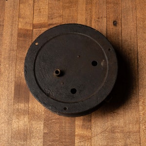 May include: A round, black metal clock face with a brass knob and three holes. The clock face is on a wooden surface.