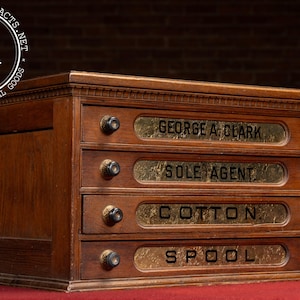 May include: A vintage wooden cabinet with four drawers. Each drawer has a brass nameplate with text: "GEORGE A. CLARK", "SOLE AGENT", "COTTON", and "SPOOL". The cabinet has dark wooden knobs and is set against a dark background.