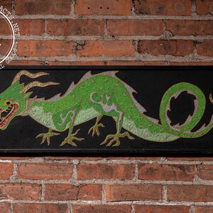 May include: A rectangular black framed wall art piece featuring a green dragon with gold accents. The dragon is depicted in a side profile with its mouth open, revealing red details. The artwork is mounted on a brick wall.