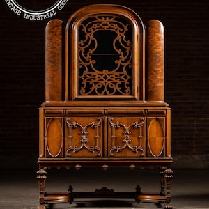 May include: An antique wooden cabinet with intricate carvings and a dark interior. The cabinet features a central arched section with decorative fretwork, flanked by two columns. The base has two doors with ornate details and sits on carved legs.