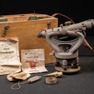 May include: A grey metal transit level with adjustment knobs, a wooden storage case, a manual titled Manual of instructions for the USE and CARE of the Level and Transit-Level, a parts list, a plumb bob, and a spool of string with a metal weight.
