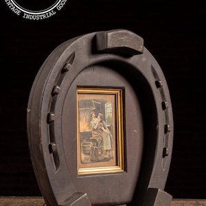 May include: A dark brown horseshoe-shaped frame with a rectangular picture of a blacksmith scene. The frame has a vintage industrial aesthetic, with visible nail heads and a rustic finish. The frame is standing on a wooden surface.
