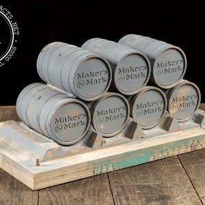 May include: A display of seven small, gray wooden barrels stacked on a wooden base. Each barrel has the words "Maker's Mark" printed on the front. The base is painted gray and has some green paint splatters.
