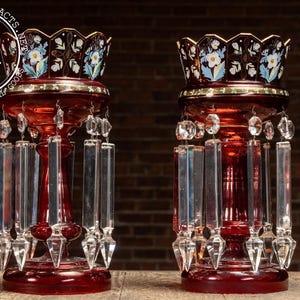 May include: Two antique ruby red glass candle holders with crystal prism accents. The tops are decorated with floral designs in white, blue, and gold. The candle holders are displayed on a wooden surface against a brick wall.
