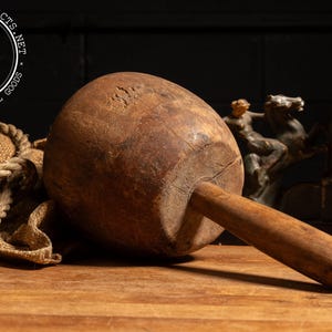 May include: A large, antique wooden mallet with a rounded head and a long handle. The mallet is made of aged, weathered wood, showing signs of wear. A vintage book and a sculpture are in the background. The mallet is likely used for woodworking or construction.