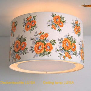 Könnte beinhalten: Eine zylindrische Deckenleuchte mit weißem Hintergrund und einem wiederkehrenden Blumenmuster aus orangefarbenen Mohnblumen und kleinen blauen Blüten. Die Lampe strahlt oben und unten warmes Licht aus und trägt den Text Deckenleuchte LUISA.
