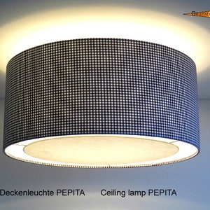 Peut inclure: Un plafonnier cylindrique avec un abat-jour à motif vichy noir et blanc. La lampe est éclairée de l'intérieur, diffusant une lueur chaleureuse. Le texte "Ceiling lamp PEPITA" est visible.