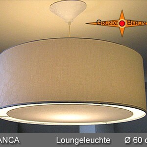 Può includere: Una lampada a sospensione cilindrica con paralume in tessuto beige e diffusore inferiore circolare. L'apparecchio è sospeso a un rosone bianco. Il testo GRUZDZ BERLIN è visibile in alto a destra, e BLANCA Loungeleuchte 60 cm in basso.