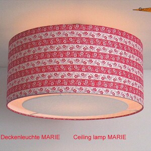 Puede incluir: Una lámpara de techo cilíndrica con una pantalla de tela que presenta bandas horizontales con motivos florales rojos y blancos. La lámpara emite un brillo cálido e incluye el texto Deckenleuchte MARIE y Ceiling lamp MARIE en la parte inferior.