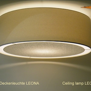 May include: A beige ceiling lamp with a round, fabric shade. The lamp is lit and casts a warm glow. The text "Deckenleuchte LEONA" and "Ceiling lamp LEONA" is visible below the lamp.