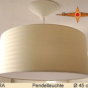Könnte beinhalten: Eine beige zylindrische Pendelleuchte mit strukturiertem Stoffschirm und passendem konischem Deckenbaldachin. Die Leuchte ist mit DORA beschriftet und enthält ein Logo von GRUZDZ BERLIN. Der Durchmesser der Leuchte beträgt 45 cm.