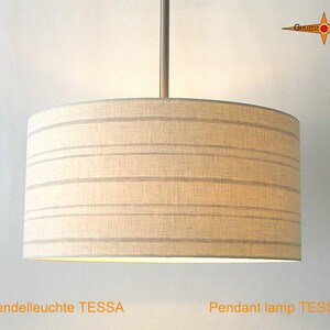 May include: A cylindrical pendant lamp with a beige fabric shade featuring thin horizontal grey stripes. The lamp is suspended by a central metal rod. The text GRUZDZ BERLIN is visible in the top right corner, and the text Pendant lamp TESSA is at the bottom.