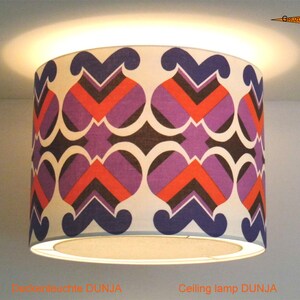Op de afbeelding: Een witte plafondlampkap met een geometrisch patroon in tinten paars, oranje en zwart. De lampkap is gelabeld "Plafondlamp DUNJA".