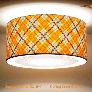 Può includere: Una lampada da soffitto cilindrica con un vivace motivo a quadri arancione e giallo. Il paralume presenta un design geometrico ripetuto con sottili linee incrociate rosso scuro e bianche. La lampada emette una luce brillante e riporta la scritta Ceiling lamp SUNNY.