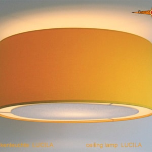 Könnte beinhalten: Eine gelbe Deckenleuchte mit einem weißen Innenschirm. Die Lampe ist mit "Deckenleuchte LUCILA" und "ceiling lamp LUCILA" beschriftet.