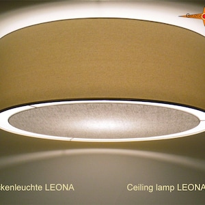 Könnte beinhalten: Eine beige Deckenleuchte mit einem runden, stoffbezogenen Schirm. Die Lampe ist eingeschaltet und strahlt ein warmes Licht aus. Der Text "Deckenleuchte LEONA" und "Ceiling lamp LEONA" ist unterhalb der Lampe sichtbar.