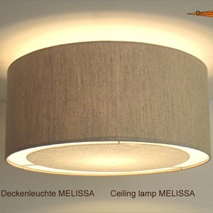 Könnte beinhalten: Eine zylindrische Deckenleuchte namens Melissa mit einem strukturierten beigen Stoffschirm. Die Leuchte strahlt ein warmes, diffuses Licht an den oberen und unteren Rändern aus, das die Deckenfläche beleuchtet und die runde Bodenplatte erhellt.