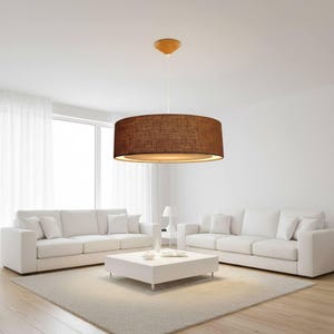 Può includere: Una lampada a sospensione marrone a tamburo è appesa al soffitto in un moderno soggiorno. L'apparecchio ha un paralume cilindrico con un aspetto strutturato. La stanza è dotata di divani bianchi, un tavolino bianco e un tappeto colorato.