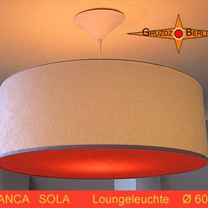 May include: A cylindrical pendant light fitting featuring a cream-coloured fabric exterior and a vibrant orange interior lining. The lamp is suspended from a white ceiling canopy. The text GRUZDZ BERLIN and BLANCA SOLA Loungeleuchte Ø 60 appear.