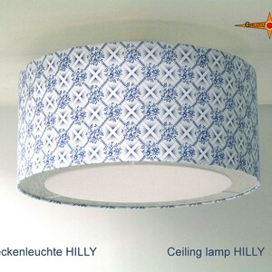 May include: A white ceiling lamp with a blue and white patterned shade. The shade has a geometric pattern of blue flowers and white lines. The text "Deckenleuchte HILLY" and "Ceiling lamp HILLY" is visible below the lamp.