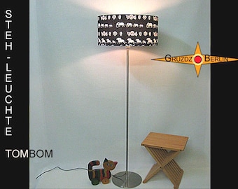 Peut inclure: Un lampadaire avec un abat-jour cylindrique à motif d'éléphants noirs et blancs, un pied métallique fin et une base circulaire. Le texte sur l'image indique STEH-LEUCHTE, TOMBOM et GRUZDZ BERLIN. Un tabouret pliant en bois et un jouet chat sont présents.