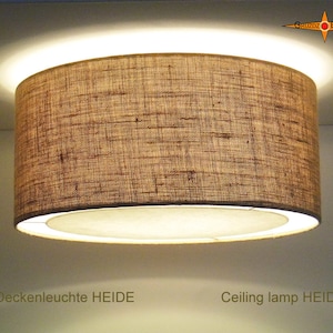 Può includere: Una lampada da soffitto cilindrica marrone con un paralume in tessuto. La lampada è accesa ed emette una luce calda. Il testo "Deckenleuchte HEIDE" e "Ceiling lamp HEIDE" è visibile sotto la lampada.