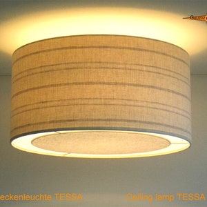 Könnte beinhalten: Eine beige Deckenleuchte mit einem gestreiften Stoffschirm. Die Lampe ist eingeschaltet und strahlt ein warmes Licht. Der Text "Deckenleuchte TESSA" ist unter der Lampe sichtbar.