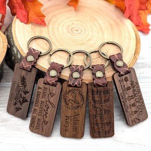 May include: A collection of personalized wooden keychains with leather straps and metal rings. Each keychain is engraved with text, including names, initials, and inspirational quotes. The keychains are brown and feature a variety of fonts and designs.
