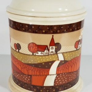 May include: A ceramic cookie jar with a white lid and a colorful patchwork design featuring a small town with houses and trees. The jar has a brown base with a white rim.