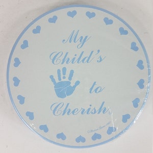 May include: A white round ceramic plate with a blue border and blue hearts around the edge. The plate has a blue handprint in the center and the text "My Child's to Cherish" in blue cursive writing. The text "Perine Jones 1981" is in smaller blue writing at the bottom.