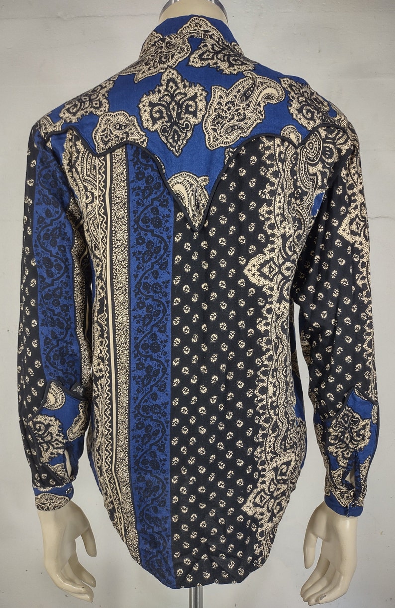 May include: A blue, black, and cream paisley patterned long-sleeve shirt with a pointed yoke in the back.