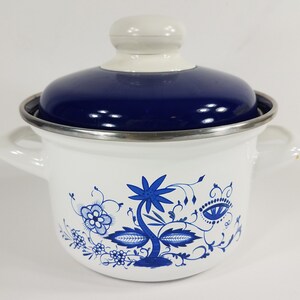 May include: White enamel pot with a blue lid and floral design. The pot has two side handles and a silver rim. The blue floral pattern decorates the white exterior. The lid has a white knob.