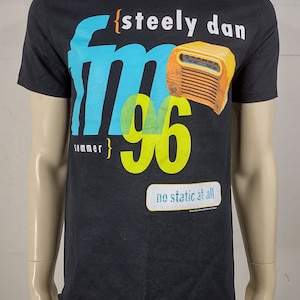 May include: Black t-shirt with a graphic design featuring the words "steely dan fm 96 summer" in white, blue, and yellow. An illustration of a vintage radio is also included. The text "no static at all" is below the radio.