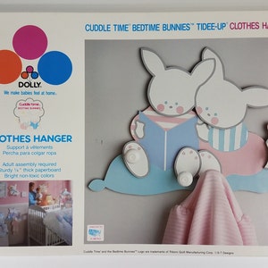 May include: A white and blue children's clothes hanger with two bunnies reading a book. The hanger is made of sturdy paperboard and has a pink, blue, and white color scheme. The text on the box reads "Cuddle Time Bedtime Bunnies™ Tidee-Up Clothes Hanger" and "No. 426".