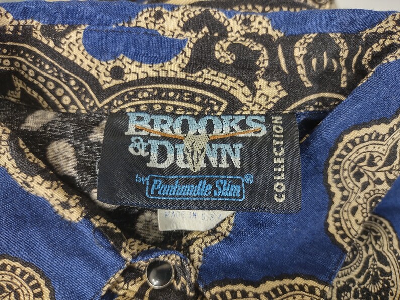 May include: A blue and tan paisley patterned shirt with a black label that reads "Brooks & Dunn by Panhandle Slim" and "Collection" in white text.