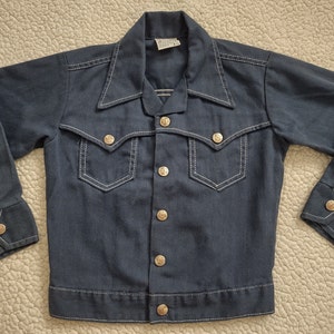 May include: A dark blue denim jacket with white stitching and gold buttons. The jacket has two chest pockets and two front pockets.