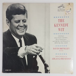 May include: A black and white photo of John F. Kennedy smiling while speaking into a microphone. The album cover features the title "The Kennedy Wit" with the text "Selections from famous Speeches, Press Conferences and Off-the-Cuff Remarks - Edited by Bill Adler". The album cover also features the text "David Brinkley Narrator" and "Spoken Introduction by Adlai E. Stevenson".