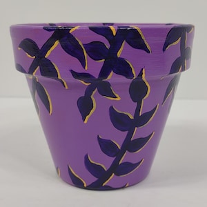 May include: Purple terracotta flower pot with a hand-painted design of dark purple leaves with yellow outlines.