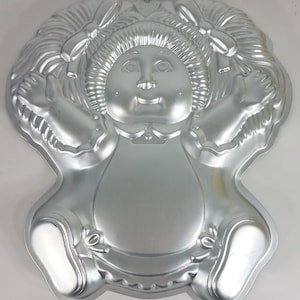 May include: Silver-toned metal cake pan shaped like a doll with pigtails and a dress. The doll has a round face with a smile and large eyes.